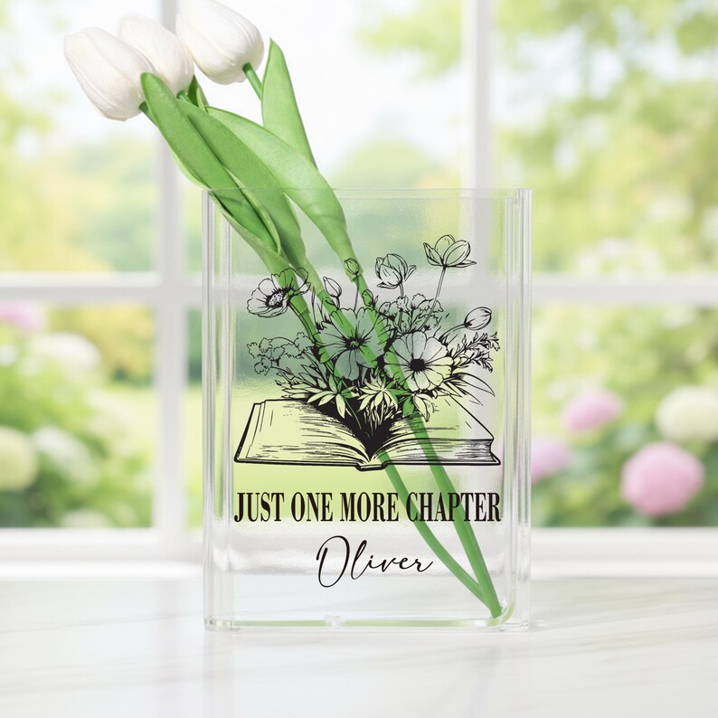 May include: Clear acrylic vase with a floral design and the text "JUST ONE MORE CHAPTER Oliver." The vase holds white tulips with green stems and leaves. The floral design features an open book with flowers.