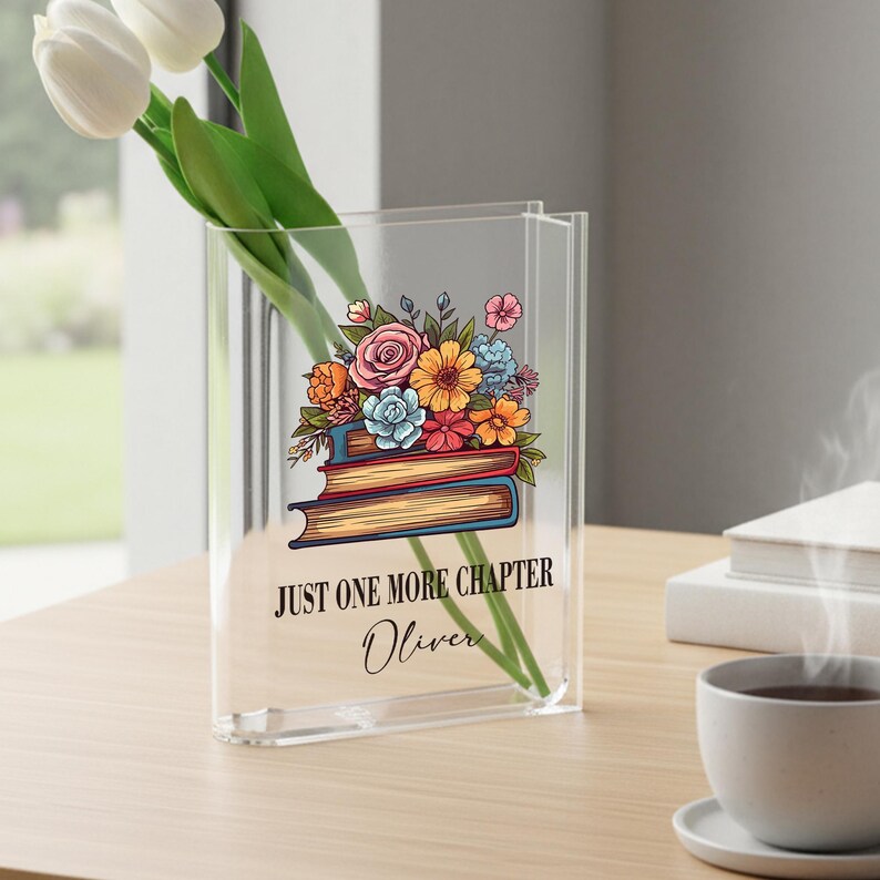 May include: Clear acrylic vase with a floral and book design. The vase features a colourful bouquet of flowers atop two books, with the text "JUST ONE MORE CHAPTER Oliver" printed below. White tulips are placed inside the vase.