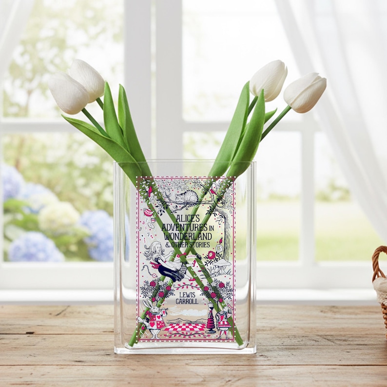 May include: A clear glass vase with a rectangular shape, featuring a decorative panel with illustrations and text from "Alice's Adventures in Wonderland & Other Stories" by Lewis Carroll. The vase holds three white tulips with green stems.