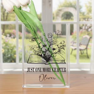 May include: Clear acrylic vase with a floral design and the text "JUST ONE MORE CHAPTER Oliver". The vase holds white tulips with green stems and leaves. The floral design features an open book with flowers.