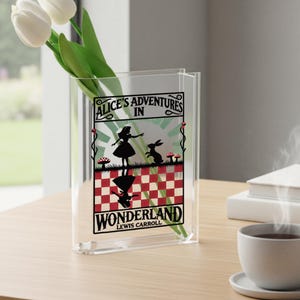 May include: Clear acrylic vase with the text "Alice's Adventures in Wonderland" and a silhouette of Alice and the rabbit. The vase holds white tulips and is on a wooden table.