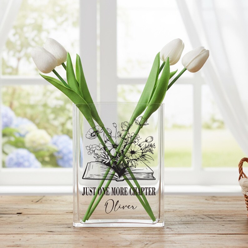 May include: A clear glass vase with a floral and book design, holding three white tulips with green stems. The vase has the text "JUST ONE MORE CHAPTER" and the name "Oliver". The vase is on a wooden surface.