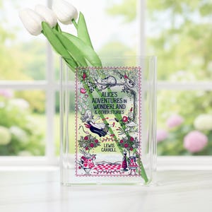 May include: A clear acrylic vase with a colorful illustration of Alice in Wonderland. The illustration features the text "Alice's Adventures in Wonderland & Other Stories" and includes images of Alice, a cat, and other characters. White tulips are inside the vase.
