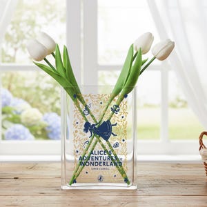 May include: A clear glass vase with an "Alice's Adventures in Wonderland" design in gold and navy blue. The vase holds three white tulips with green stems. The vase is on a wooden surface in front of a window.