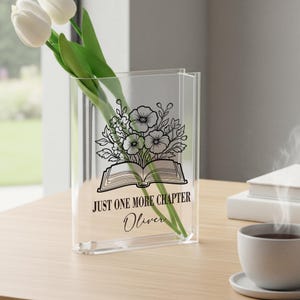 May include: Clear acrylic vase featuring a floral and book design, with the text "JUST ONE MORE CHAPTER" and the name "Oliver". The vase holds white tulips with green stems. The vase is on a wooden table.
