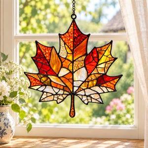 May include: A stained glass suncatcher shaped like a maple leaf, featuring a mosaic of red, orange, yellow, and clear glass segments held together by black metal cames. The leaf hangs from a black metal chain against a window background.