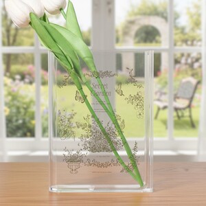 Pode incluir: Vaso retangular transparente com um design floral e de borboletas, e as palavras "Secret Garden". O vaso contém tulipas brancas com caules e folhas verdes. O vaso está sobre uma superfície de madeira.