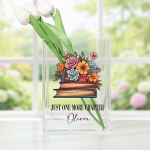 May include: A clear acrylic vase with a floral design and the text "JUST ONE MORE CHAPTER" and the name "Oliver". The vase holds white tulips. The floral design features a stack of books with colorful flowers.