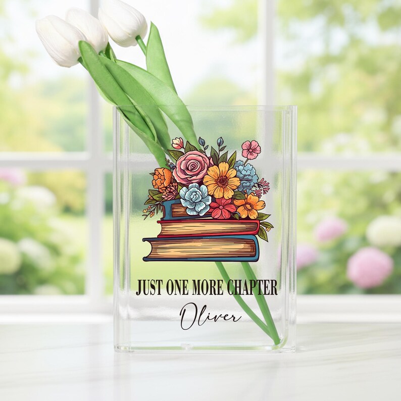 May include: A clear acrylic vase with a floral design and the text "JUST ONE MORE CHAPTER" and the name "Oliver". The vase holds white tulips. The floral design features a stack of books with colorful flowers.