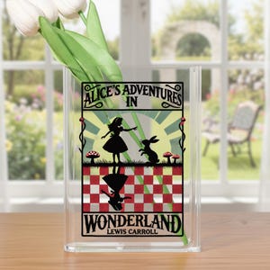 May include: Clear acrylic vase with a black graphic of Alice in Wonderland. The graphic includes the text "Alice's Adventures in Wonderland" and illustrations of Alice, a rabbit, and mushrooms. White tulips are inside the vase.