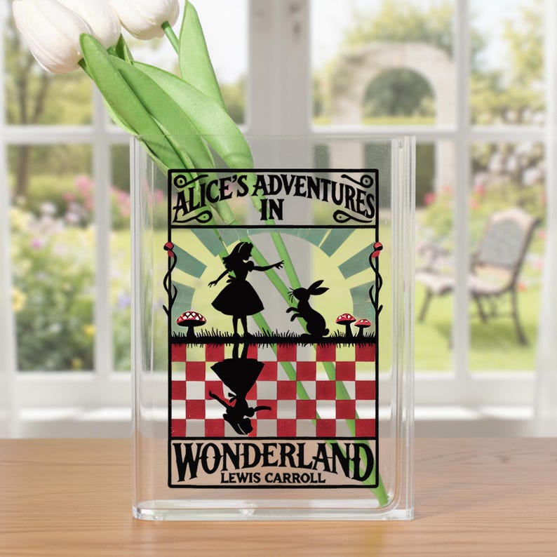 May include: Clear acrylic vase with a black graphic of Alice in Wonderland. The graphic includes the text "Alice's Adventures in Wonderland" and illustrations of Alice, a rabbit, and mushrooms. White tulips are inside the vase.