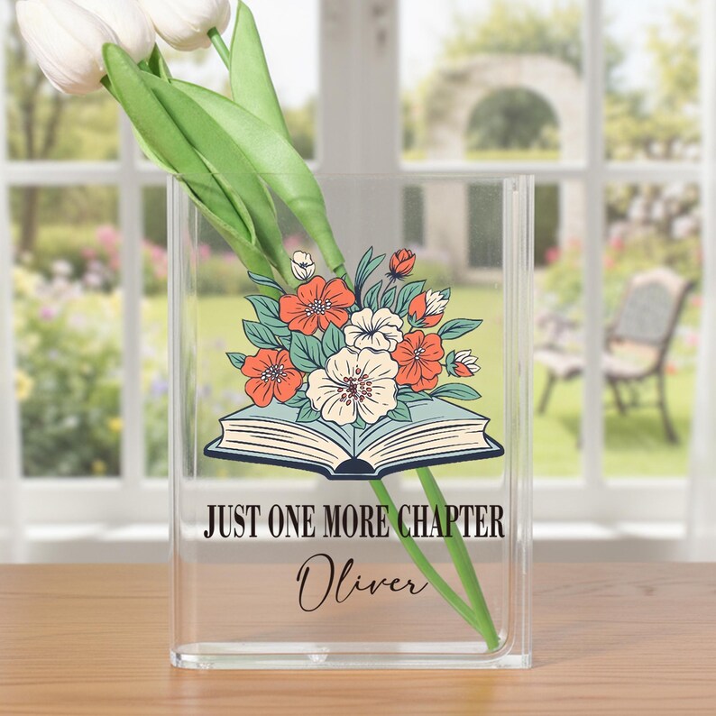 May include: Clear acrylic vase with a floral design featuring orange and white flowers, and the text "JUST ONE MORE CHAPTER" and the name "Oliver". The vase holds white tulips.