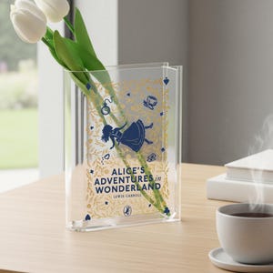 May include: Clear acrylic vase featuring the text "Alice's Adventures in Wonderland" with illustrations of Alice, a pocket watch, and a teacup. The vase holds white tulips. A decorative item for book lovers.