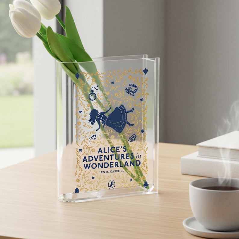 May include: Clear acrylic vase featuring the text "Alice's Adventures in Wonderland" with illustrations of Alice, a pocket watch, and a teacup. The vase holds white tulips. A decorative item for book lovers.