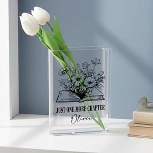 May include: A clear acrylic vase shaped like a book, with the text "JUST ONE MORE CHAPTER" and the name "Oliver". The vase holds white tulips and a black and white floral illustration. The vase is on a white surface.