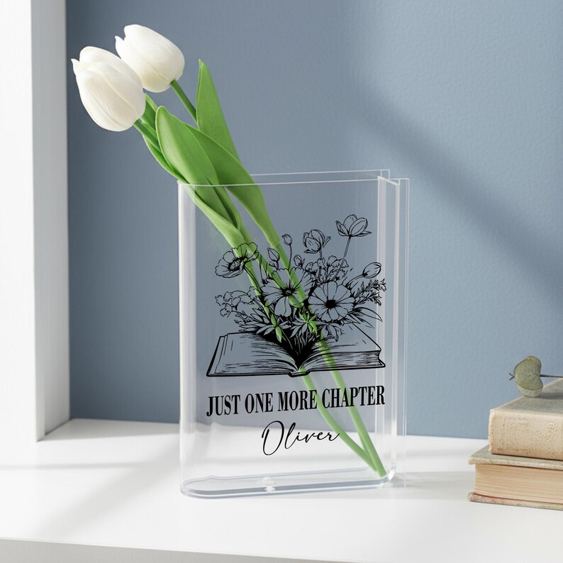 May include: A clear acrylic vase shaped like a book, with the text "JUST ONE MORE CHAPTER" and the name "Oliver". The vase holds white tulips and a black and white floral illustration. The vase is on a white surface.