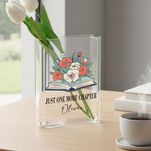 May include: Clear acrylic vase with a floral design featuring orange and white flowers, and the text "JUST ONE MORE CHAPTER Oliver". The vase holds white tulips. The vase is on a wooden surface.