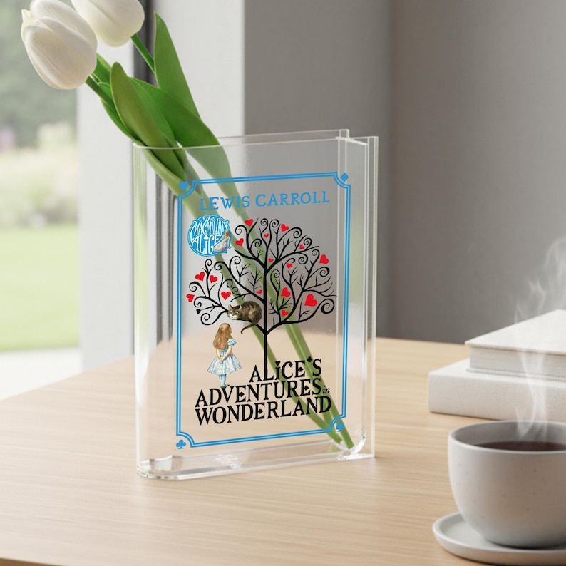 May include: Clear acrylic vase with a design from "Alice's Adventures in Wonderland." The design features a tree with red heart-shaped leaves, a cat, and Alice. The vase holds white tulips. Text includes "Lewis Carroll" and "Alice's Adventures in Wonderland."