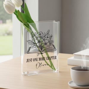 May include: Clear acrylic vase featuring a floral and book design, with the text "JUST ONE MORE CHAPTER" and the name "Oliver". White tulips are displayed inside the vase. The vase is on a wooden table.