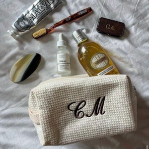 Puede incluir: Un surtido de artículos de tocador y productos de belleza, que incluyen un tubo de pasta de dientes Marvis, un cepillo de dientes, una botella de aceite de ducha y un sérum para el cuidado de la piel. También hay un neceser de color crema con las iniciales "CM" bordadas, junto con una pequeña caja con las iniciales "C.A."