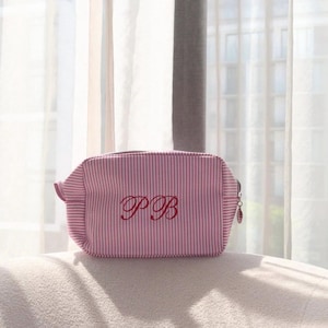 May include: A pink and white striped cosmetic bag with the embroidered initials "PB" in a script font. The bag has a zipper closure and a small loop on the side. The bag is sitting on a white surface.