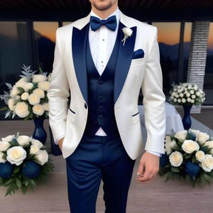 May include: An ivory suit with navy blue detailing, including the lapels, waistcoat, and trousers. The ensemble features a navy bow tie, a white shirt, and a pocket square. A white flower adorns the lapel. The image depicts formal attire, suitable for a wedding.