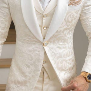 May include: Cream-coloured formal suit with a delicate floral design. The suit comprises a jacket with a shawl collar, a waistcoat, and matching trousers. Gold buttons and a pocket square provide detail. A gold watch is visible.