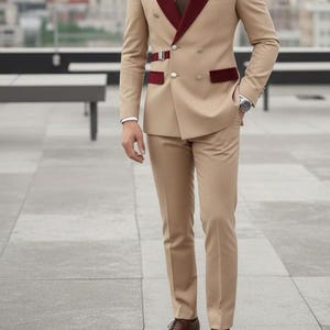 May include: A tan double-breasted suit with burgundy velvet lapels and accents. The suit features a matching tie, pocket square, and brown leather shoes. The image includes the text "HussainbyAnas".