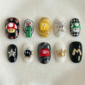 K&ouml;nnte beinhalten: Zehn k&uuml;nstliche Fingern&auml;gel mit verschiedenen Designs, die von dem Videospiel Super Mario inspiriert sind. Die N&auml;gel zeigen ikonische Elemente wie einen Pilz, einen Fragezeichenblock, einen Stern und Marios Hut in verschiedenen Farben.