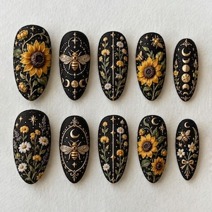 May include: A set of ten almond-shaped press-on nails featuring a matte black base with raised 3D designs including yellow sunflowers, white daisies, gold bees, celestial moon phases, and star accents arranged in intricate botanical patterns.