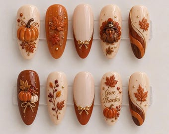 May include: A set of ten almond-shaped press-on nails featuring Thanksgiving themes. Designs include 3D pumpkins, maple leaves, a turkey, gold bows, and the text Give Thanks. The color palette consists of cream, orange, brown, and metallic gold accents.