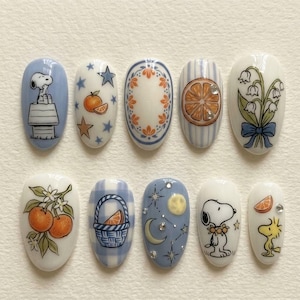 Puede incluir: Diez dise&ntilde;os de u&ntilde;as ovaladas con ilustraciones. Los dise&ntilde;os presentan a Snoopy, naranjas, patrones florales y motivos celestiales en azul, blanco, naranja y verde. Algunas u&ntilde;as tienen un acabado brillante, mostrando detalles.