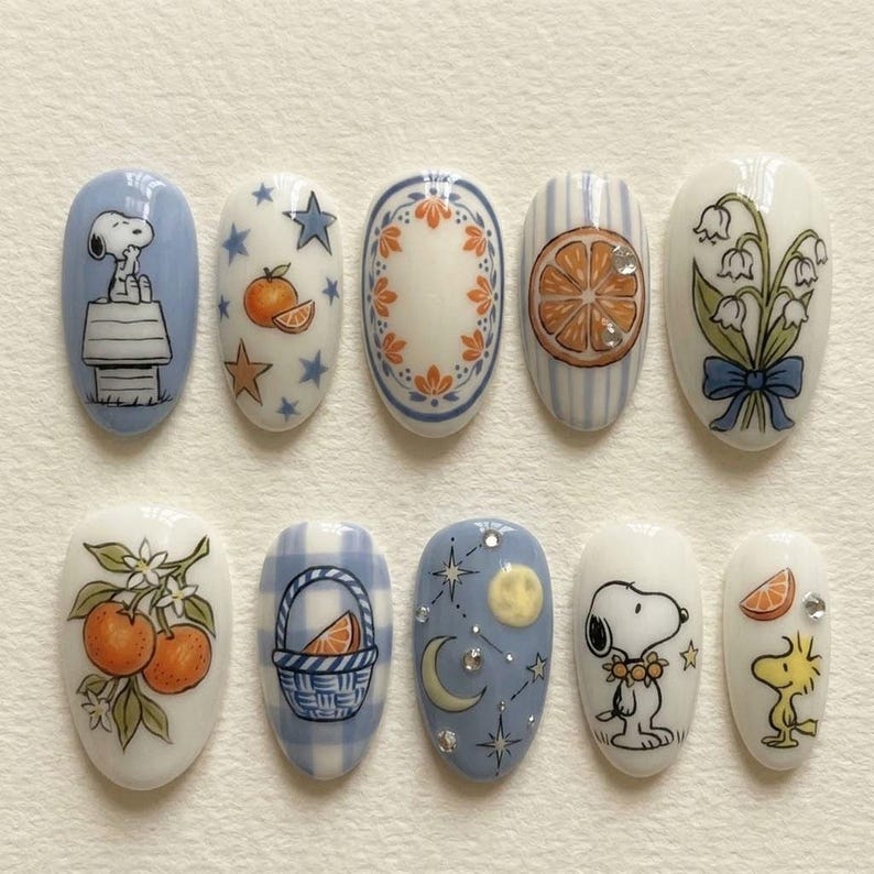 Puede incluir: Diez dise&ntilde;os de u&ntilde;as ovaladas con ilustraciones. Los dise&ntilde;os presentan a Snoopy, naranjas, patrones florales y motivos celestiales en azul, blanco, naranja y verde. Algunas u&ntilde;as tienen un acabado brillante, mostrando detalles.