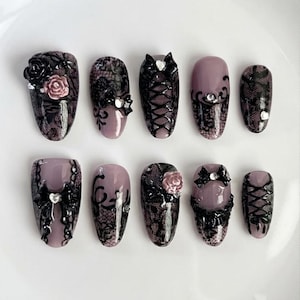 May include: A set of ten press-on nails featuring a mauve base with black lace patterns, 3D black bows, small pink rose accents, corset-style lacing details, and clear rhinestones. The nails have a glossy finish and varied decorative designs.