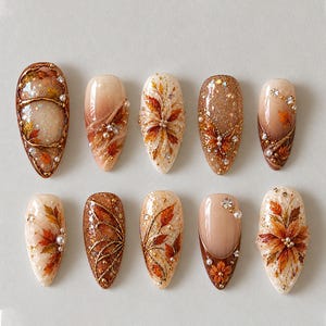 May include: A set of ten almond-shaped press-on nails featuring autumn-themed designs. The nails display a colour palette of cream, beige, and deep brown, decorated with painted orange and red maple leaves, gold glitter, small white pearls, and clear rhinestones.