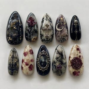 May include: A set of ten almond-shaped press-on nails featuring intricate designs including a dark rose, a silver cross, a crescent moon with stars, a gothic castle in a frame, a wax seal, and ornate black filigree patterns on marble and cream bases.