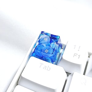 May include: A custom keycap with a blue floral design encased in clear resin. The keycap is on a white keyboard, with the "Tab" key visible below. The keycap has a unique, artistic appearance.