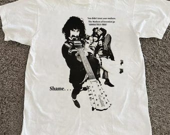 May include: White t-shirt featuring a black and white graphic of a musician playing a guitar. The graphic includes the text "Shame..." and "You didn't trust your mothers. The Mothers of Invention go 'ABSOLUTELY FREE'".