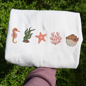 May include: White tote bag featuring embroidered sea creatures: a seahorse, seaweed, starfish, coral, and a pufferfish. The illustrations are in shades of orange, green, and brown. The bag is held against a background of green foliage.