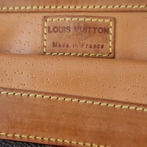 Puede incluir: Primer plano de una cartera de cuero marrón con el logotipo de Louis Vuitton. La cartera presenta una superficie texturizada, costuras amarillas y una etiqueta que dice "LOUIS VUITTON PARIS Made in France."