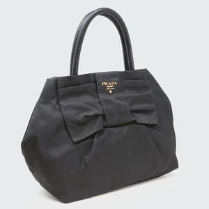 以下が含まれることがあります： 大きなリボンと革のハンドルが付いた黒いハンドバッグ。バッグには、リボンに金色のロゴが付いています。バッグはトートスタイルです。