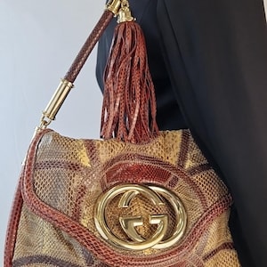 May include: A luxury handbag with a snakeskin pattern in shades of brown, tan, and red. The bag features a gold-tone Gucci logo and hardware, a braided shoulder strap, and a tassel. The bag is a shoulder bag.