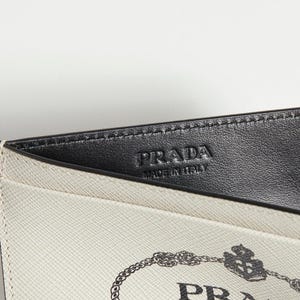 以下が含まれることがあります： プラダのカードケースのクローズアップ。カードケースは、クリーム色の外側と黒革の内側が特徴です。前面にはプラダのロゴが見えます。「PRADA MADE IN ITALY」の文字が黒革にエンボス加工されています。