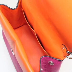 Puede incluir: Primer plano de un bolso magenta con interior naranja. El bolso tiene un cierre de solapa con un cierre plateado. El interior tiene dos bolsillos y está forrado con cuero naranja.