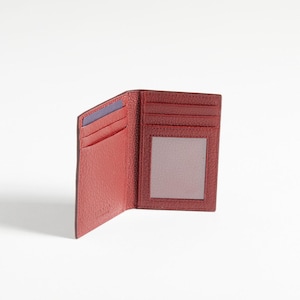 Può includere: Portafoglio in pelle rosso aperto con più fessure per carte e una finestra trasparente per documenti d'identità. Il portafoglio è esposto su uno sfondo bianco, evidenziando la sua superficie strutturata e i dettagli interni. È visibile una carta blu.