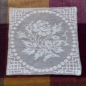 May include: A square white crochet doily featuring a central floral motif with detailed rose petals and leaves. The piece is crafted with a fine grid-like mesh pattern, creating a delicate openwork design that highlights the intricate needlework.