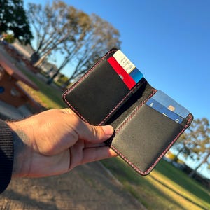 May include: Black leather wallet with pink stitching, open to display credit cards. The wallet is held against a backdrop of a park with trees and a blue sky.