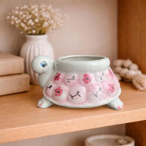 May include: A ceramic turtle-shaped planter with a light green body and a pink shell decorated with red flowers. The planter has a wide opening and is sitting on a wooden shelf. The turtle has blue eyes and is a decorative item.