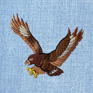 May include: Embroidered patch of a brown eagle in flight with outstretched wings and yellow talons. The eagle is detailed with shades of brown and white, set against a light blue denim background.