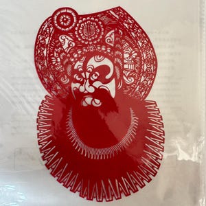 May include: A red paper cut-out depicting a traditional Chinese opera figure. The figure has a detailed headdress and beard, with intricate patterns and designs. The cut-out is likely used for decoration or as a cultural craft.